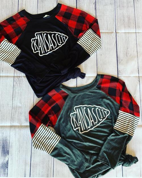 Two stylish Kansas City graphic tees with plaid and striped sleeves, laid out on a wooden surface.