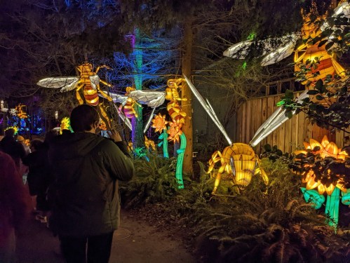 A person takes a photo of colorful illuminated bees and flowers in a festive outdoor display at night.