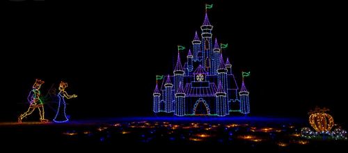 A brightly lit castle made of colorful lights, with two figures walking nearby and a glowing pumpkin in the foreground.