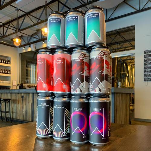 A stack of colorful beer cans on a wooden table, with a brewery backdrop featuring various beer styles.