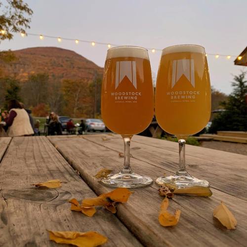 Two glasses of beer on a wooden table, surrounded by autumn leaves, with a mountain and string lights in the background.