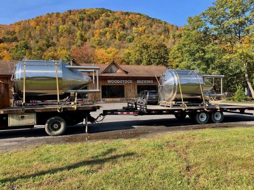 Two large stainless steel brewing tanks on a trailer in front of a brewery, surrounded by autumn foliage.