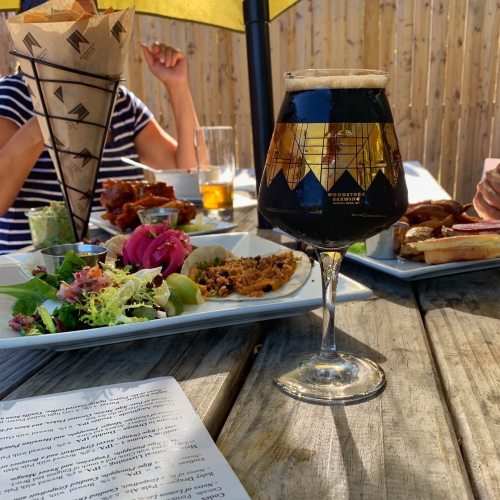 A glass of dark beer sits on a wooden table, surrounded by plates of food and a sunny outdoor setting.