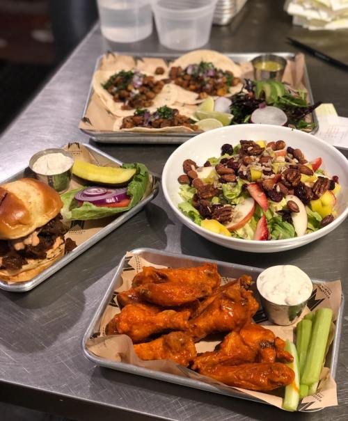 A spread of food including chicken wings, tacos, a salad with nuts and fruit, and a sandwich with sides on metal trays.