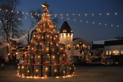 A festive Christmas tree made of stacked barrels, adorned with lights and decorations, set against a twilight sky.