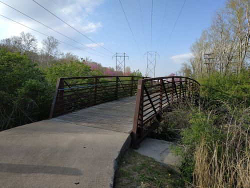 A metal bridge over a small path, surrounded by greenery and power lines in the background under a clear sky.