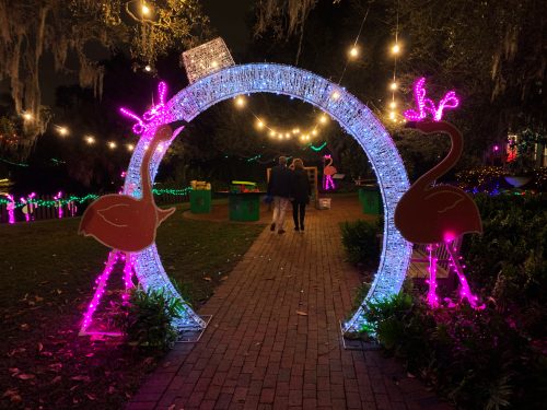 A glowing archway with pink flamingos leads to a festive pathway, illuminated by string lights and surrounded by greenery.