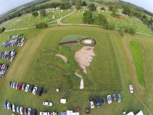 Aerial view of a large grass mural depicting a pilot's face, surrounded by cars and a cemetery in the background.