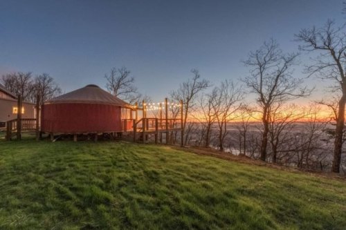 A cozy yurt on a hillside at sunset, surrounded by trees and overlooking a valley. Warm lights adorn the deck.