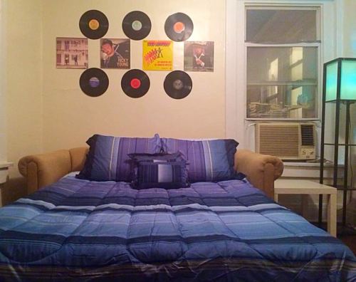 A cozy bedroom with a blue bedspread, a couch, and vinyl records on the wall above a window and air conditioner.