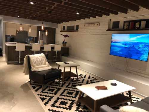 Modern basement living area with a TV, two coffee tables, a patterned rug, and a bar with stools in the background.