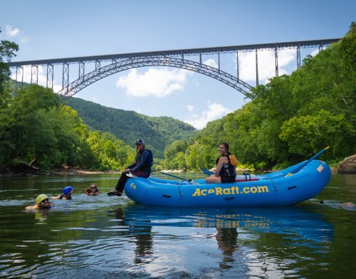 A group of people rafting in a river under a large bridge, surrounded by lush green hills and a clear blue sky.