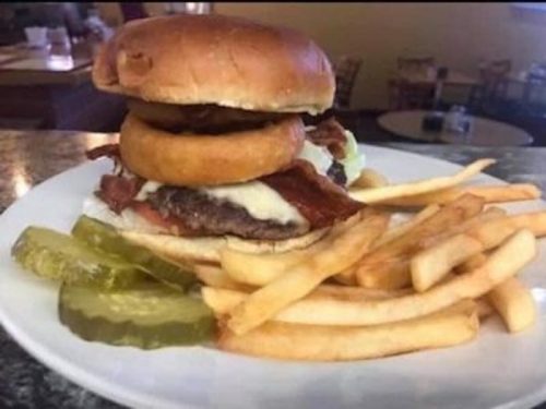 A hearty burger topped with bacon and onion rings, served with fries and pickles on the side.