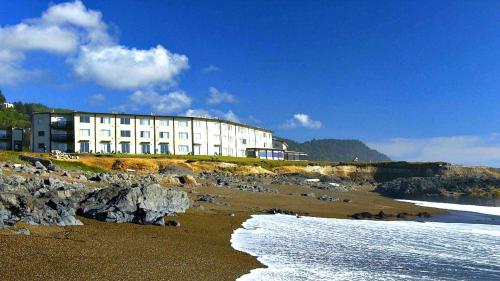 A coastal hotel sits near a rocky beach under a blue sky with scattered clouds and gentle waves lapping at the shore.