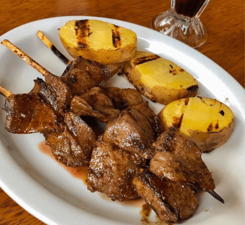 Grilled skewers of meat served with charred potato halves on a white plate. A drink is visible in the background.