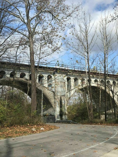 A stone bridge arches over a road, surrounded by trees and autumn leaves under a partly cloudy sky.