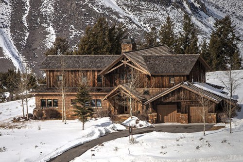 A rustic wooden house surrounded by snow and mountains, featuring a stone chimney and a winding driveway.