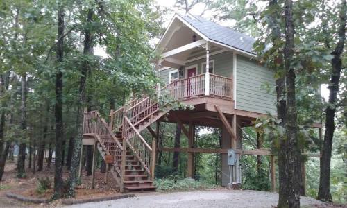 A small green house on stilts surrounded by trees, featuring a staircase leading up to a porch.