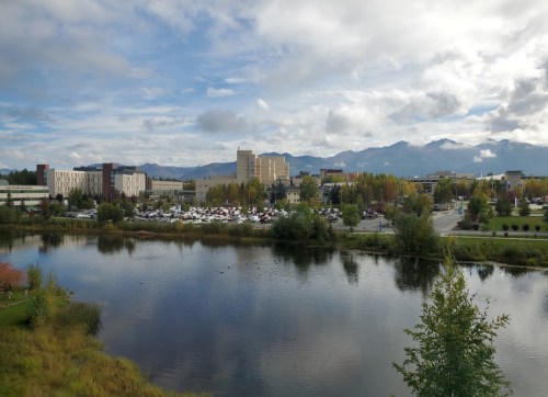A scenic view of a campus by a lake, with mountains in the background and autumn foliage.
