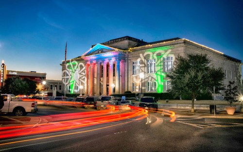 Historic building illuminated with colorful lights at dusk, surrounded by moving car lights and festive decorations.
