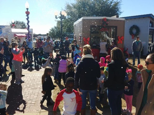 A festive crowd gathers outdoors, enjoying a snowy scene with children playing and holiday decorations in the background.
