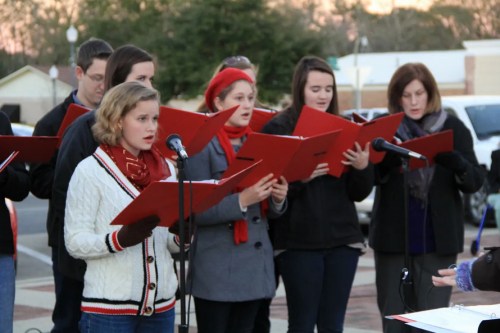 A group of singers in winter attire holds red folders while performing outdoors, with microphones in front of them.