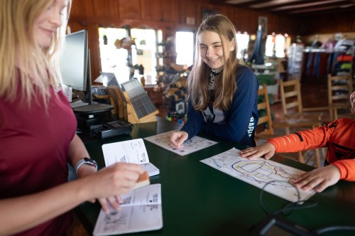 A young girl smiles at a counter while an adult stamps a passport, with another child pointing at a map.