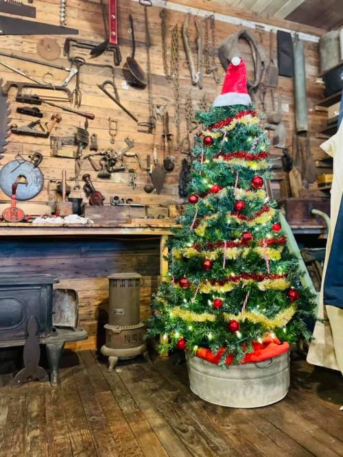 A decorated Christmas tree with a red hat stands in a rustic workshop filled with tools and wooden walls.