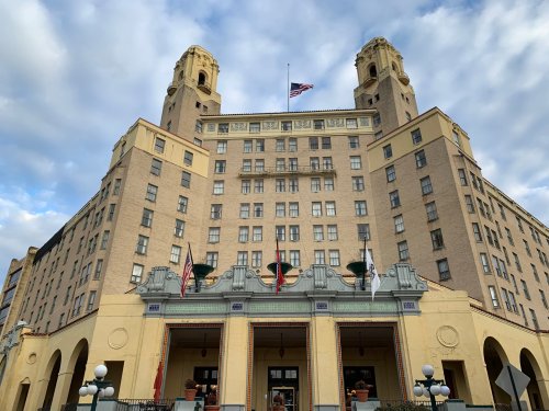 Historic hotel building with two towers, flags, and a grand entrance, set against a cloudy sky.