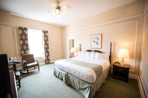 A cozy hotel room featuring a large bed, armchair, table, and window with patterned curtains. Soft lighting enhances the ambiance.