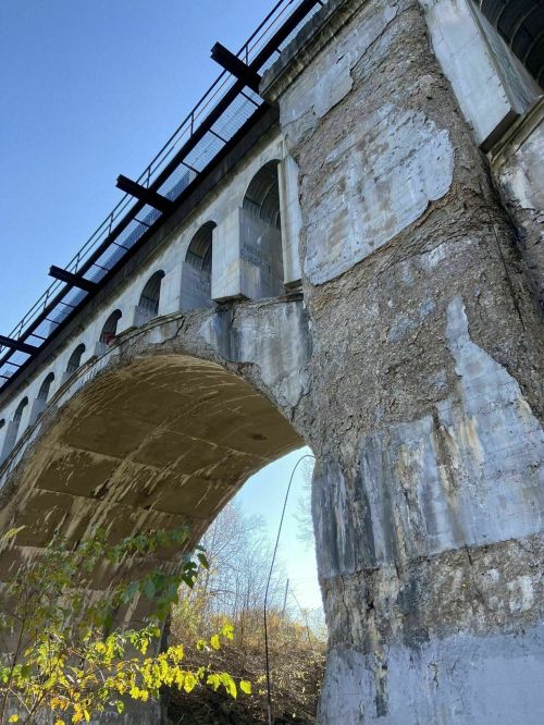 A close-up view of a weathered stone bridge with arches, under a clear blue sky and surrounded by greenery.