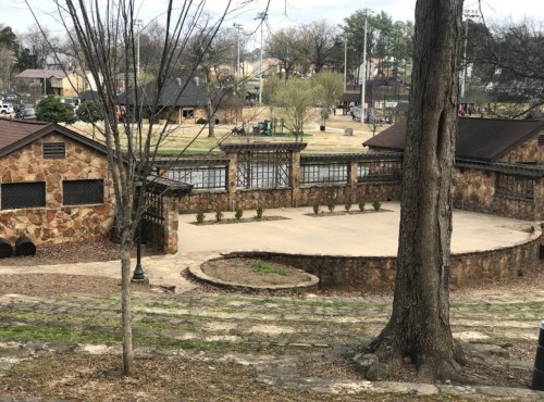 A stone amphitheater surrounded by trees, with a grassy area and buildings in the background.