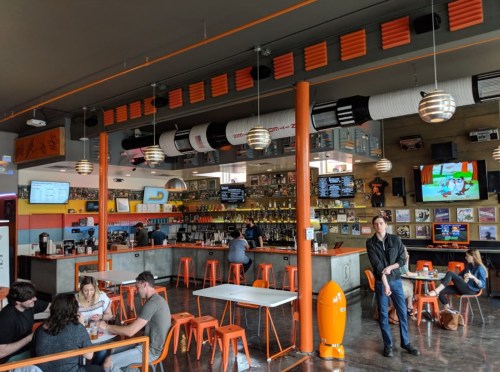 A vibrant bar with orange accents, featuring patrons at tables, a long counter, and a TV displaying sports.