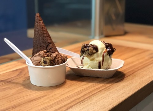 A chocolate ice cream cone and a bowl of vanilla ice cream topped with chocolate sauce and cookie pieces on a wooden surface.