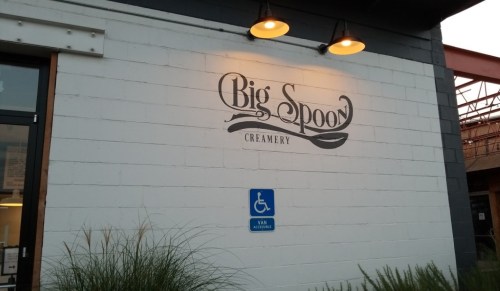 Sign for "Big Spoon Creamery" on a building wall, with outdoor lighting and a wheelchair-accessible symbol nearby.