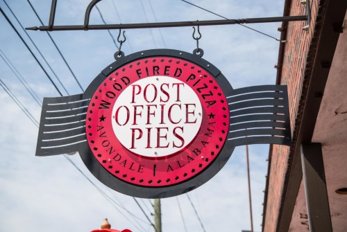 Sign for "Post Office Pies" featuring wood-fired pizza, located in Avondale, Alabama, against a blue sky.