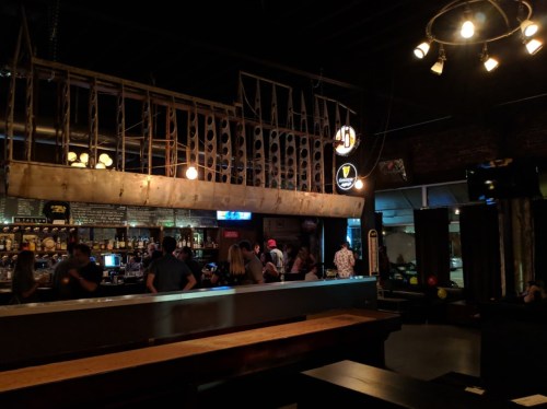 A dimly lit bar with a rustic design, featuring a long counter and patrons socializing in the background.
