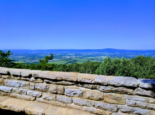 A panoramic view of a lush green valley under a clear blue sky, seen from a stone wall viewpoint.