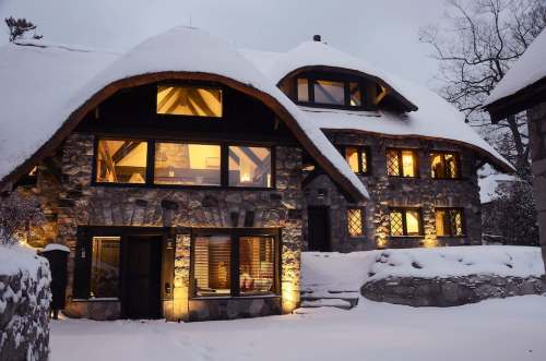 A cozy stone house with large windows, illuminated at night, surrounded by snow-covered ground and trees.