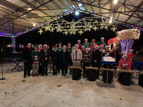 A festive group of musicians in holiday attire poses under colorful lights and decorations at an outdoor event.