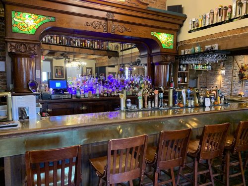A wooden bar with colorful stained glass, lined with stools and a variety of liquor bottles on display.