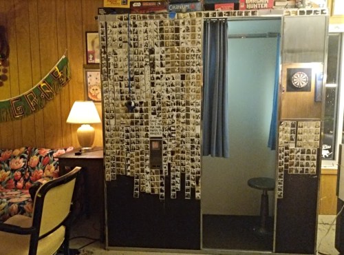 A vintage photo booth covered in black-and-white photos, with a curtain, dartboard, and cozy seating nearby.