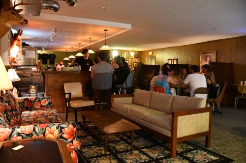A cozy bar scene with a floral couch, wooden tables, and patrons socializing in a warmly lit, retro atmosphere.
