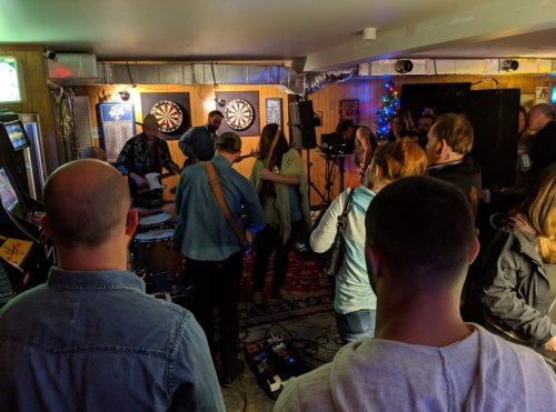 A lively indoor music performance with a crowd enjoying the show, surrounded by dartboards and festive decorations.