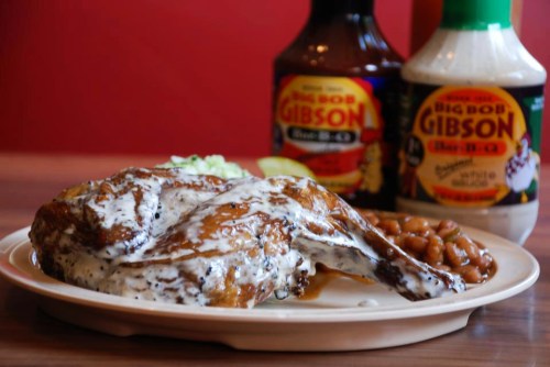A plate of grilled chicken drizzled with white sauce, served with baked beans and barbecue sauces in the background.
