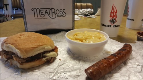 A pulled pork sandwich, a side of macaroni and cheese, and a sausage link on a table with a Meat Boss sign.