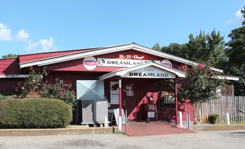 Exterior of Dreamland BBQ restaurant, featuring a red building, signage, and a clear blue sky.