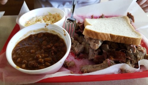 A red tray with pulled pork, a slice of bread, coleslaw, and a bowl of baked beans.