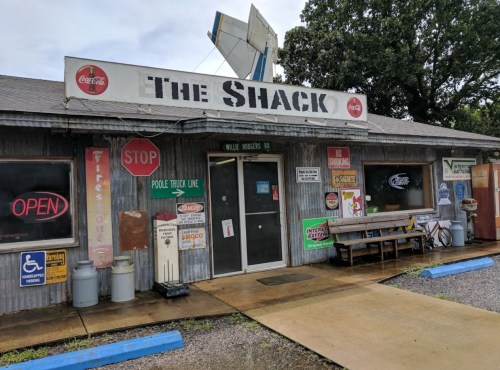 A rustic building with a sign reading "The Shack," featuring various vintage signs and an open sign in front.