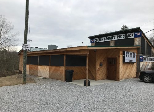 Exterior of Cooter Brown's Rib Shack, featuring wooden siding, a gravel parking area, and a sign for extra parking in back.